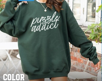 May include: Forest green sweatshirt with the text "puzzle addict" in white script. The sweatshirt has long sleeves and a crew neck. The words "Fast Shipping & More Colors" are in a blue box in the upper left corner. The words "Color Forest Green" are at the bottom.