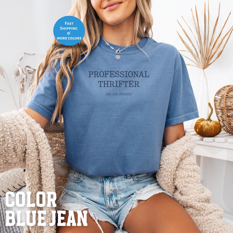 May include: Blue jean colored t-shirt with the text "PROFESSIONAL THRIFTER (do not disturb)". The shirt has short sleeves and a crew neck. A round blue sticker with the words "FAST SHIPPING & MORE COLORS" is in the upper left corner.