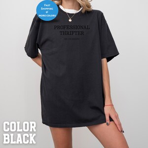 May include: Black t-shirt with the text "PROFESSIONAL THRIFTER" and "(do not disturb)" printed in black. The shirt has a round neck and short sleeves. A blue circle with the words "FAST SHIPPING & MORE COLORS" is in the upper left corner.
