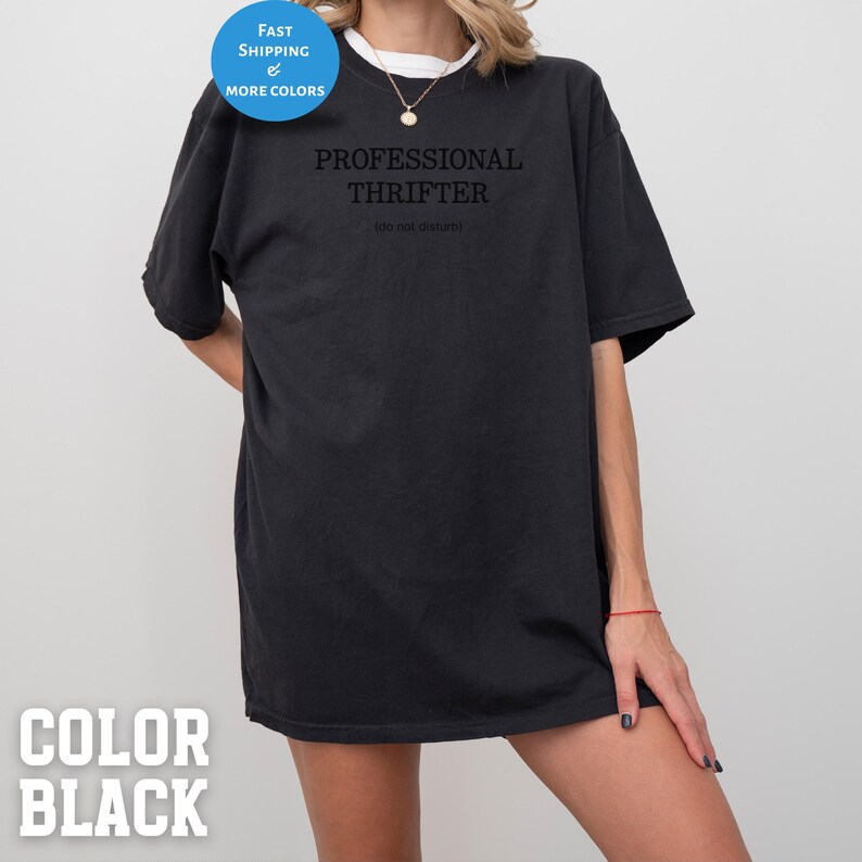 May include: Black t-shirt with the text "PROFESSIONAL THRIFTER" and "(do not disturb)" printed in black. The shirt has a round neck and short sleeves. A blue circle with the words "FAST SHIPPING & MORE COLORS" is in the upper left corner.