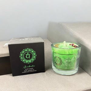 Op de afbeelding: Een groene kaars in een glazen pot met de tekst "Zarya Anahata Heart Chakra" en een zwarte doos met dezelfde tekst en een groen chakrasymbool. De kaars heeft gedroogde bloemen en kruiden bovenop.