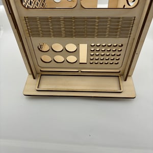 Può includere: Un modello in legno di una cabina telefonica in stile retrò con un design dettagliato. La cabina ha una piccola finestra, una porta e una serie di fori e fessure sul davanti. Il modello è realizzato in legno chiaro e ha una finitura naturale.