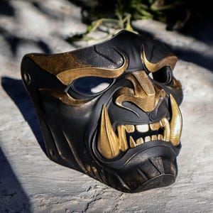 May include: A black and gold Oni mask with sharp teeth and a fierce expression. The mask has gold accents on the brow, cheeks, and fangs, and is made of a textured material. The mask is a decorative item.