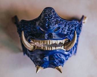 Puede incluir: Una máscara Oni azul y dorada con dientes y cuernos afilados. La máscara está hecha de plástico y tiene una superficie texturizada.