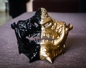 May include: A black and gold Oni mask with sharp teeth and horns. The mask is made of plastic and has a detailed design.