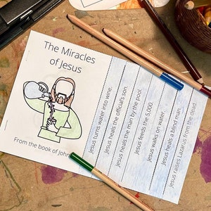 May include: A blue and white printable worksheet with the title "The Miracles of Jesus" and a list of miracles performed by Jesus. The worksheet includes a simple illustration of Jesus turning water into wine. The worksheet is on a brown surface with colored pencils and a basket of colored pencils.