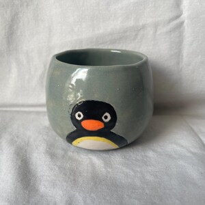 Puede incluir: Una pequeña taza de cerámica redondeada con un acabado gris liso y brillante. La parte frontal de la taza presenta un gráfico pintado a mano de un pingüino con cabeza negra, pico naranja brillante, ojos blancos y una mancha blanca en el vientre.