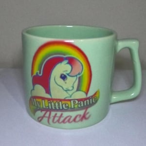 Può includere: Una tazza in ceramica verde chiaro con manico. La tazza presenta un pony dei cartoni animati con un arcobaleno e il testo "My Little Panic Attack" in rosso e giallo.
