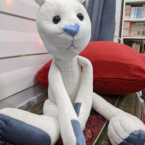 May include: White plush toy with blue accents sitting on a red pillow and a patterned rug. The toy has a long body, long arms, and long legs. The toy has a blue nose and black eyes.