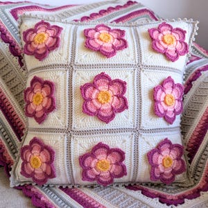 May include: A square, off-white crochet pillow with nine squares, each featuring a large, three-dimensional flower. The flowers have pink and purple petals with yellow centers. The pillow is trimmed with a decorative border and rests on a matching blanket.
