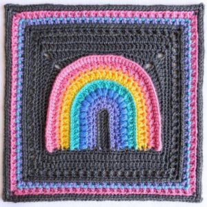 May include: A square, crocheted blanket featuring a colorful rainbow design. The rainbow arches across the center, with shades of pink, yellow, blue, and purple. The blanket has a dark gray background with a pink, blue, and purple border.