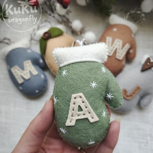 May include: A sage green felt mitten ornament featuring a white felt cuff, embroidered white snowflake details, and a cream-colored felt letter A stitched to the center. Other felt mittens in blue, tan, and gray with letters M, W, and J are visible.