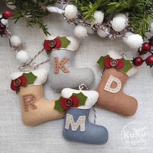 May include: Four felt Christmas stocking ornaments in light gray, tan, brown, and blue, each featuring a white cuff, a red felt holly leaf accent with a brown button, and a large letter K, R, D, or M stitched onto the front of the stocking.