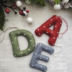 May include: Three plush felt letter ornaments in the shapes of D, A, and E. The D is olive green, the A is red, and the E is slate blue. Each letter features white embroidered star patterns and includes a hanging string attached to the top.