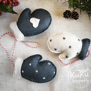 May include: Three felt mitten decorations. Two are dark grey with white trim and star or heart designs. The third is white with black star patterns. Each has a red and white string. Festive Christmas decorations.