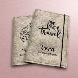 Könnte beinhalten: Zwei graue Reisepasshüllen mit schwarzer Stickerei. Die linke Hülle hat einen Globus mit dem Text "explore" und "Reise" darunter. Die rechte Hülle hat ein Herz mit einem Flugzeug und dem Text "live love travel" und "Vera Reiseunterlagen" darunter.