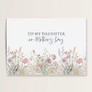 May include: A white greeting card with the text "TO MY DAUGHTER on Mother's Day" in blue script. The bottom of the card features a watercolor illustration of delicate pink, purple, and yellow flowers and greenery.