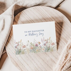 May include: A white greeting card with the text "TO MY DAUGHTER on Mother's Day" in blue script. The bottom of the card features a watercolor floral design with pink, purple, and yellow flowers. The card is on a wooden surface.
