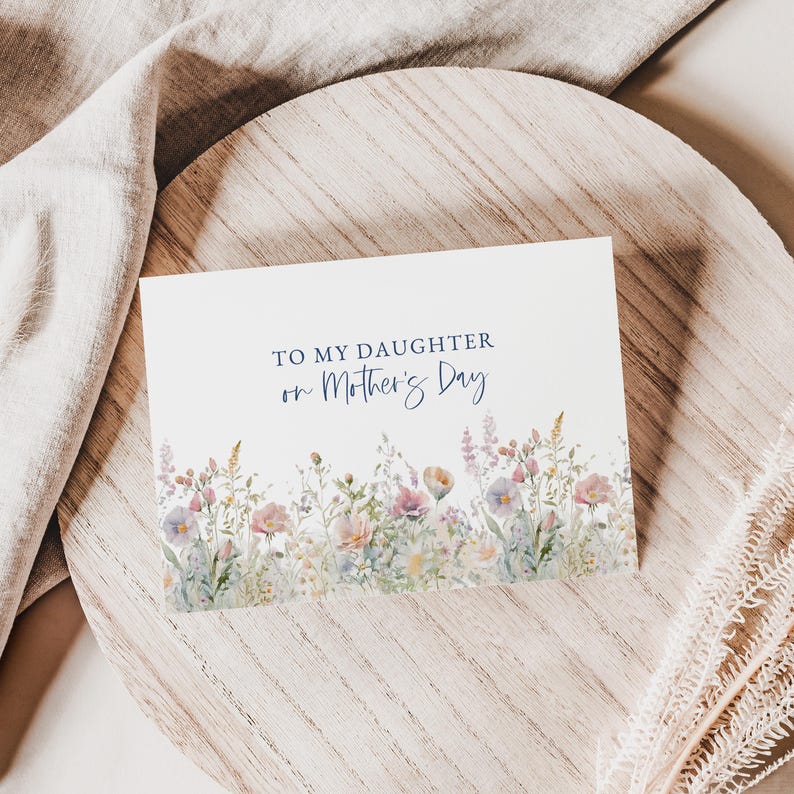 May include: A white greeting card with the text "TO MY DAUGHTER on Mother's Day" in blue script. The bottom of the card features a watercolor floral design with pink, purple, and yellow flowers. The card is on a wooden surface.