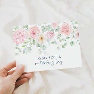 Puede incluir: Una tarjeta de felicitaci&oacute;n con un dise&ntilde;o floral con rosas rosas e hortensias, con follaje verde. La tarjeta dice "TO MY SISTER on Mother's Day" en una elegante escritura azul marino. La tarjeta se sostiene sobre un fondo blanco.