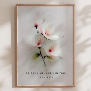 Op de afbeelding: Een ingelijste kunstdruk met een aquarel schilderij van witte bloemen met roze centra en accenten. Het kunstwerk bevat de tekst "ABIDE IN ME, AND I IN YOU. JOHN 15:4" in een eenvoudig lettertype. De lijst is licht van kleur.