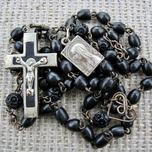 May include: An antique rosary with black beads, a silver crucifix with a figure of Jesus, and a silver pendant with a religious image. The rosary features a metal chain and decorative elements.