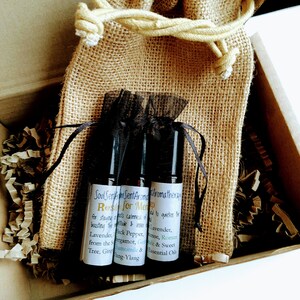 May include: Aromatherapy essential oil roller bottles in a burlap bag, inside a cardboard box. The bottles have labels with text. The bag is tied with a beige string. The box contains brown paper filler.