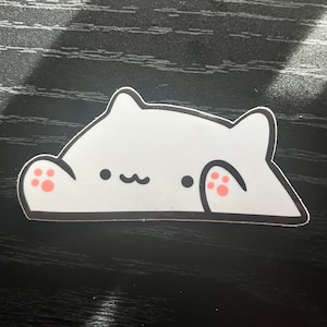 Puede incluir: Pegatina de gato blanco con huellas de patas rosas y contorno negro. El gato está tumbado con la cabeza girada hacia un lado y los ojos cerrados. La pegatina está sobre una superficie de madera oscura.