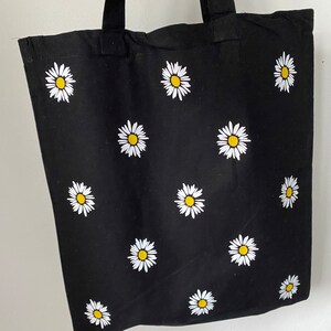 May include: Black canvas tote bag with white daisy flowers printed on it. The bag has black straps.