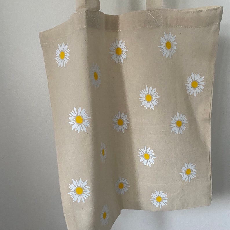 May include: A beige canvas tote bag with a white daisy pattern. The daisies have yellow centers.