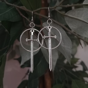 May include: Silver earrings with a sword design inside a circle. The earrings are hanging from a leafy green plant.