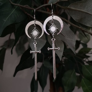 Pode incluir: Brincos de prata com um design de lua crescente e uma estrela no centro. Os brincos têm uma espada pendurada na parte inferior.