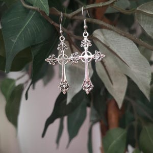 May include: A pair of silver-colored cross earrings with intricate filigree detailing. The earrings are suspended from simple hooks and are displayed against a backdrop of green leaves and branches, creating a natural contrast.