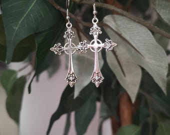 May include: A pair of silver-colored cross earrings with intricate filigree detailing. The earrings are suspended from simple hooks and are displayed against a backdrop of green leaves and branches, creating a natural contrast.
