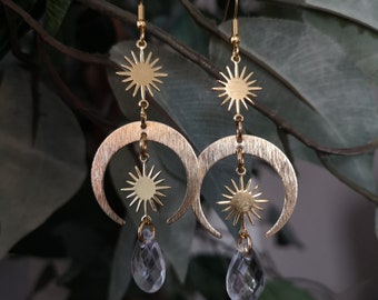 May include: A pair of gold-toned dangle earrings featuring a sunburst charm at the top, a brushed metal crescent moon in the center, a smaller sunburst charm below the moon, and a clear, faceted teardrop-shaped crystal bead at the bottom.