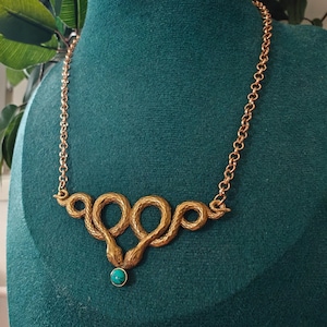 May include: A gold-toned necklace featuring a central pendant of two intertwined snakes with textured scales. The snakes meet at the bottom, where a small, round, bright turquoise stone is set. The pendant is attached to a simple gold-toned chain.