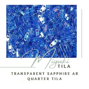 May include: A close-up image of a pile of small, transparent blue glass beads with an iridescent finish. The beads are shaped like small rectangles with two holes on each side. The beads are labeled "Miyuki Tila Transparent Sapphire AB Quarter Tila".
