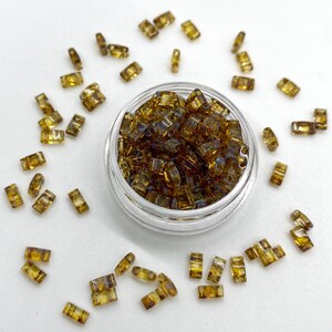 May include: A small glass jar filled with iridescent brown glass beads. The beads are rectangular in shape and have a slightly irregular texture. The jar is sitting on a white surface, and there are several loose beads scattered around it.