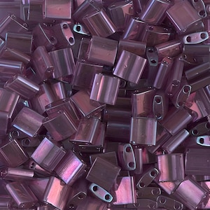 May include: A close-up shot of a pile of iridescent, rectangular beads in shades of purple and mauve. Each bead has two holes for stringing. The beads are scattered, creating a textured surface.