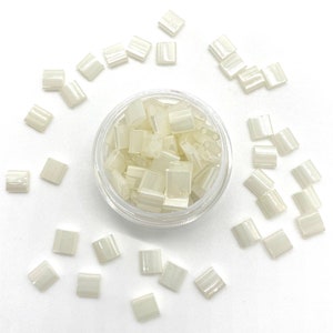 May include: A small clear container filled with white opaque square beads. The beads are scattered around the container on a white surface.