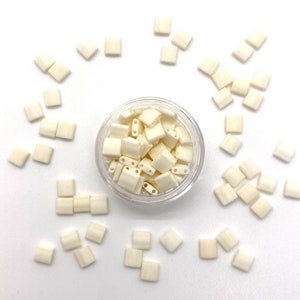 May include: A small glass jar filled with white square beads. The jar is surrounded by loose white square beads on a white background.