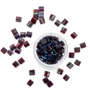 May include: A bowl filled with small, square, iridescent glass beads in shades of blue, red, and black. The beads are scattered around the bowl on a white surface.