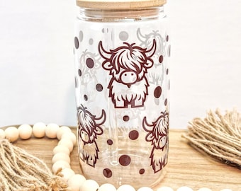 Può includere: Un bicchiere in vetro trasparente con una stampa grafica marrone scuro raffigurante tre mucche Highland e pois sparsi. Il bicchiere cilindrico include un coperchio in bambù chiaro con un foro centrale e una cannuccia in vetro dritta.