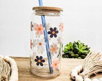 May include: A clear glass water bottle with a wooden lid and a blue reusable straw. The bottle is decorated with pink, white and black daisy stickers.