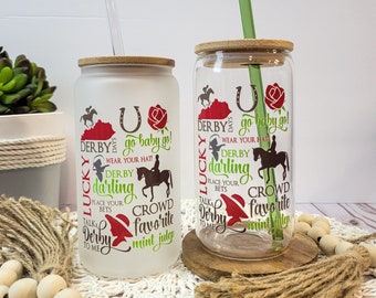 Könnte beinhalten: Zwei gefrostete Glasbecher mit einem Holzdeckel und einem Strohhalm. Die Becher haben einen weißen Hintergrund mit einer roten Rose und einem Hufeisen. Der Text auf den Bechern lautet "Derby Days", "Lucky", "Go Baby Go", "Wear Your Hat", "Derby Darling", "Place Your Bets", "Talk to Me", "Crowd Favorite", und "Mint Julep".