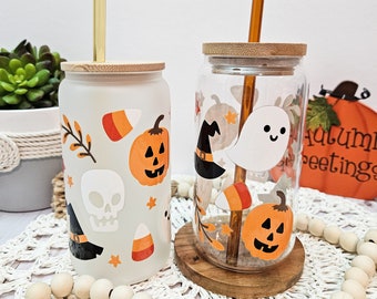 Puede incluir: Dos vasos de vidrio con tapas y pajitas, decorados con diseños de temática de Halloween. Un vaso presenta un fantasma, un sombrero de bruja y maíz dulce, mientras que el otro presenta una calavera, una calabaza y maíz dulce. Ambos vasos tienen una tapa de madera y un cuerpo de vidrio transparente.
