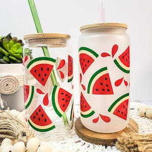 Puede incluir: Dos vasos de vidrio con diseños de impresión de sandía y tapas de bambú. Un vaso es de vidrio transparente con una pajita verde, el otro es de vidrio esmerilado con una pajita rosa. Ambos vasos están sobre una superficie de crochet blanco.