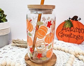 Puede incluir: Un vaso de vidrio transparente con tapa de bambú y una pajita. El vaso tiene un diseño de otoño con calabazas naranjas y hojas rojas. El texto "Autumn Greetings" es visible en una calabaza decorativa en el fondo.