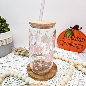 May include: A glass tumbler with a wooden lid and a straw. The tumbler has a pink and white design with pumpkins, stars, and flowers. The tumbler is sitting on a wooden coaster and a white crocheted doily. The text "Autumn Greetings" is visible in the background.