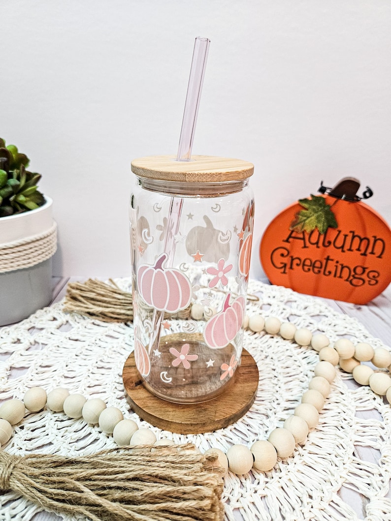 May include: A glass tumbler with a wooden lid and a straw. The tumbler has a pink and white design with pumpkins, stars, and flowers. The tumbler is sitting on a wooden coaster and a white crocheted doily. The text "Autumn Greetings" is visible in the background.